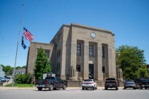 Historic Caldwell County Courthouse building with tall stone columns and a central clock, viewed from a parking area with several cars in front. Two flags, including the American flag, fly on a pole at half-staff beside the building under a clear blue sky.