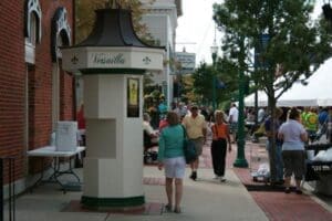 Visitors walk along a downtown sidewalk during the Versailles Poultry Days festival, with vendor booths, trees, and groups of people lining the street. A small kiosk stands in the foreground near brick storefronts, while festival activities extend into the background.