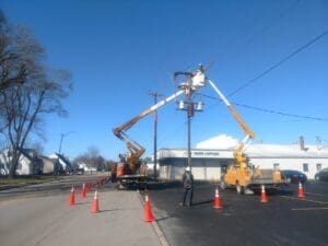 Two utility bucket trucks are positioned on either side of a utility pole as lineworkers perform maintenance on overhead power lines and electrical equipment. Orange traffic cones create a work zone along the street and parking lot, while a commercial building and leafless trees stand beneath a clear blue sky.