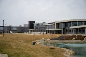 Midmark Corporation headquarters building with a modern glass-front entrance and curved facade, viewed across a landscaped campus featuring grassy areas, a small pond, and walking paths on an overcast day.